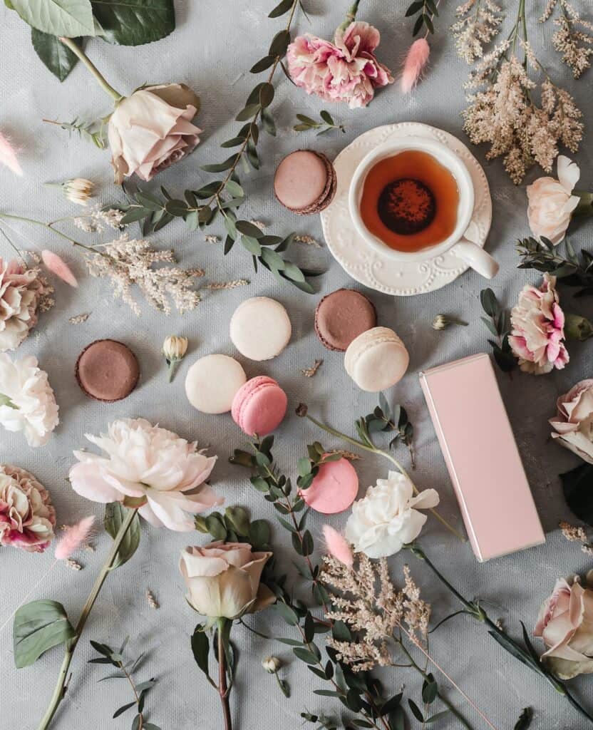 Top view of white cup with tea on saucer near various flowers and plants near macaroons on tablecloth on surface in light place